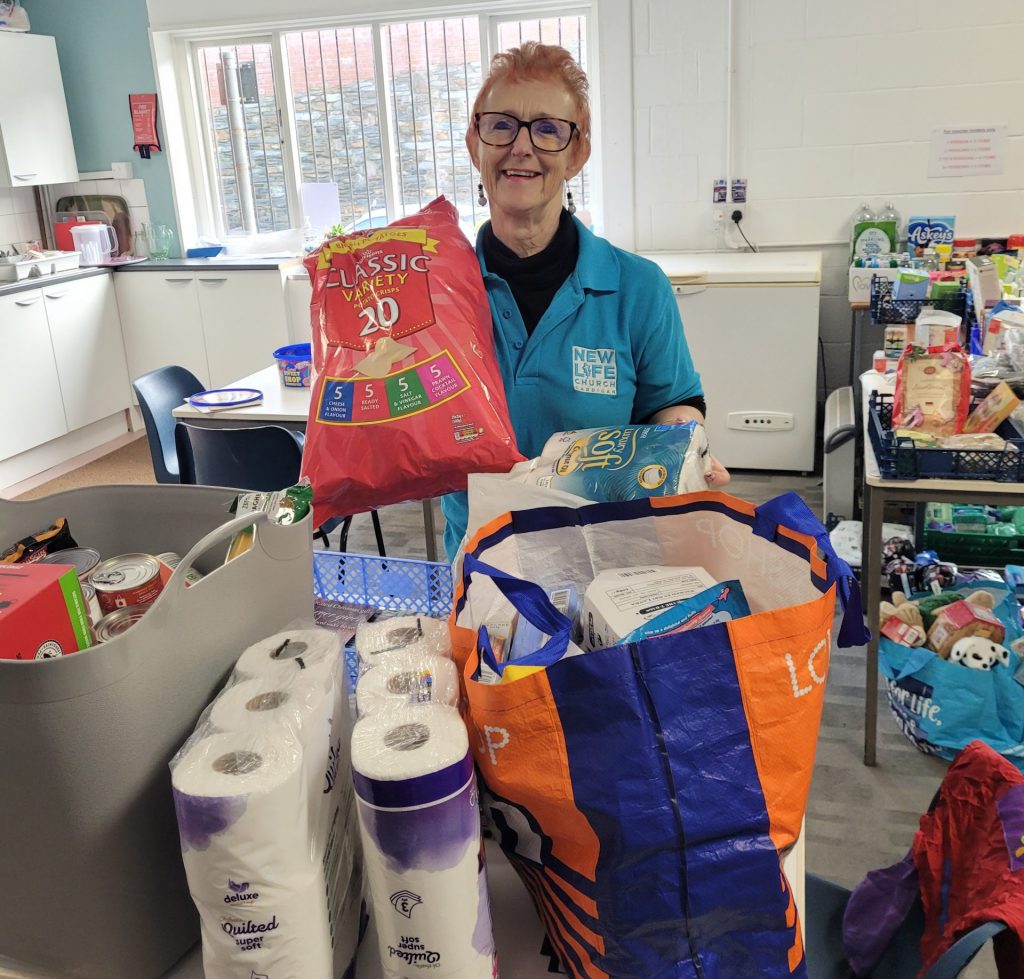 A woman in a food bank branded blue t shirt in front of a shopping bag and some toilet rolls